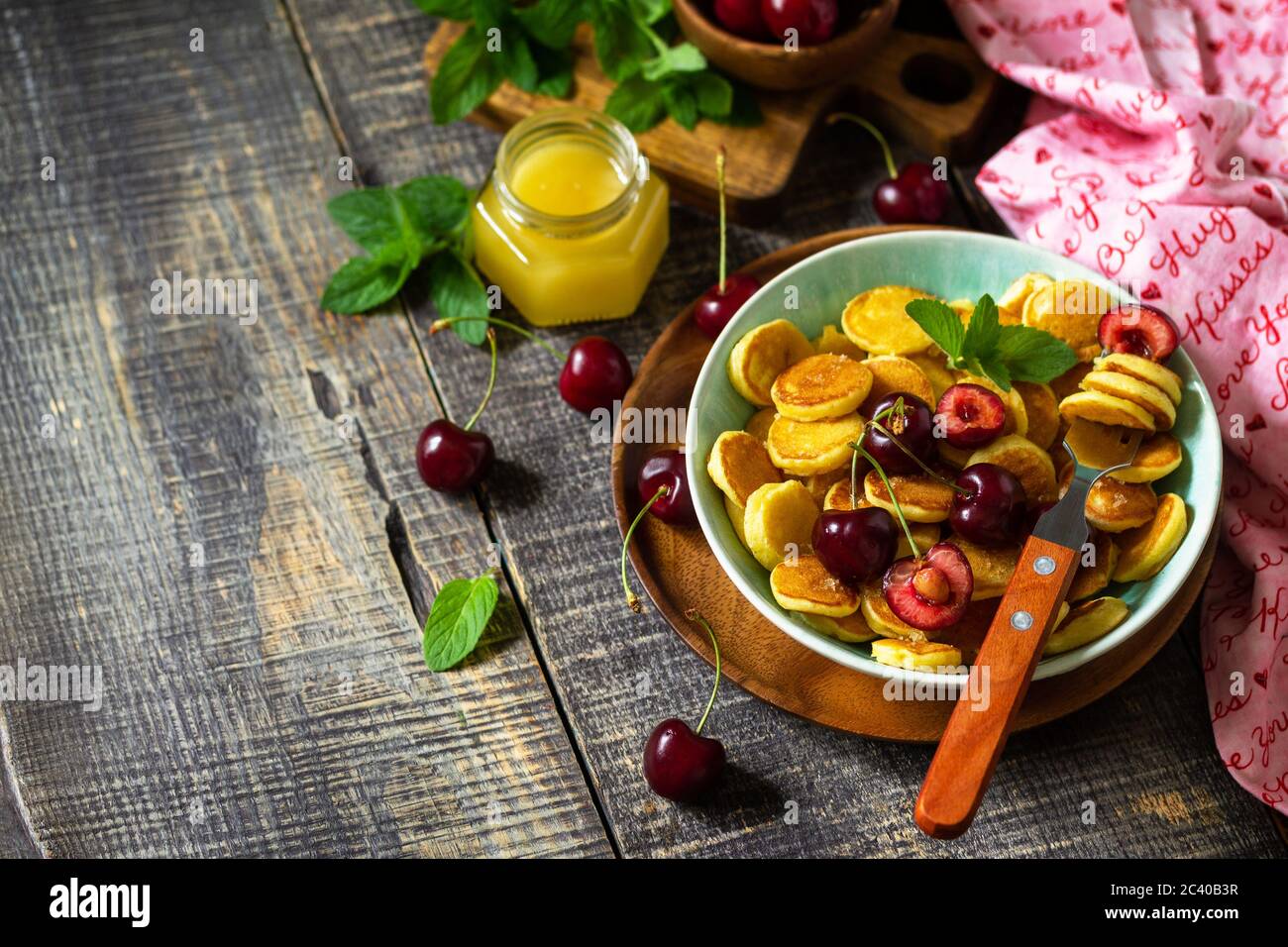 Colazione con minuscoli pancake. Mini frittelle con ciliegie e miele su rustico tavolo di legno. Spazio di copia. Foto Stock