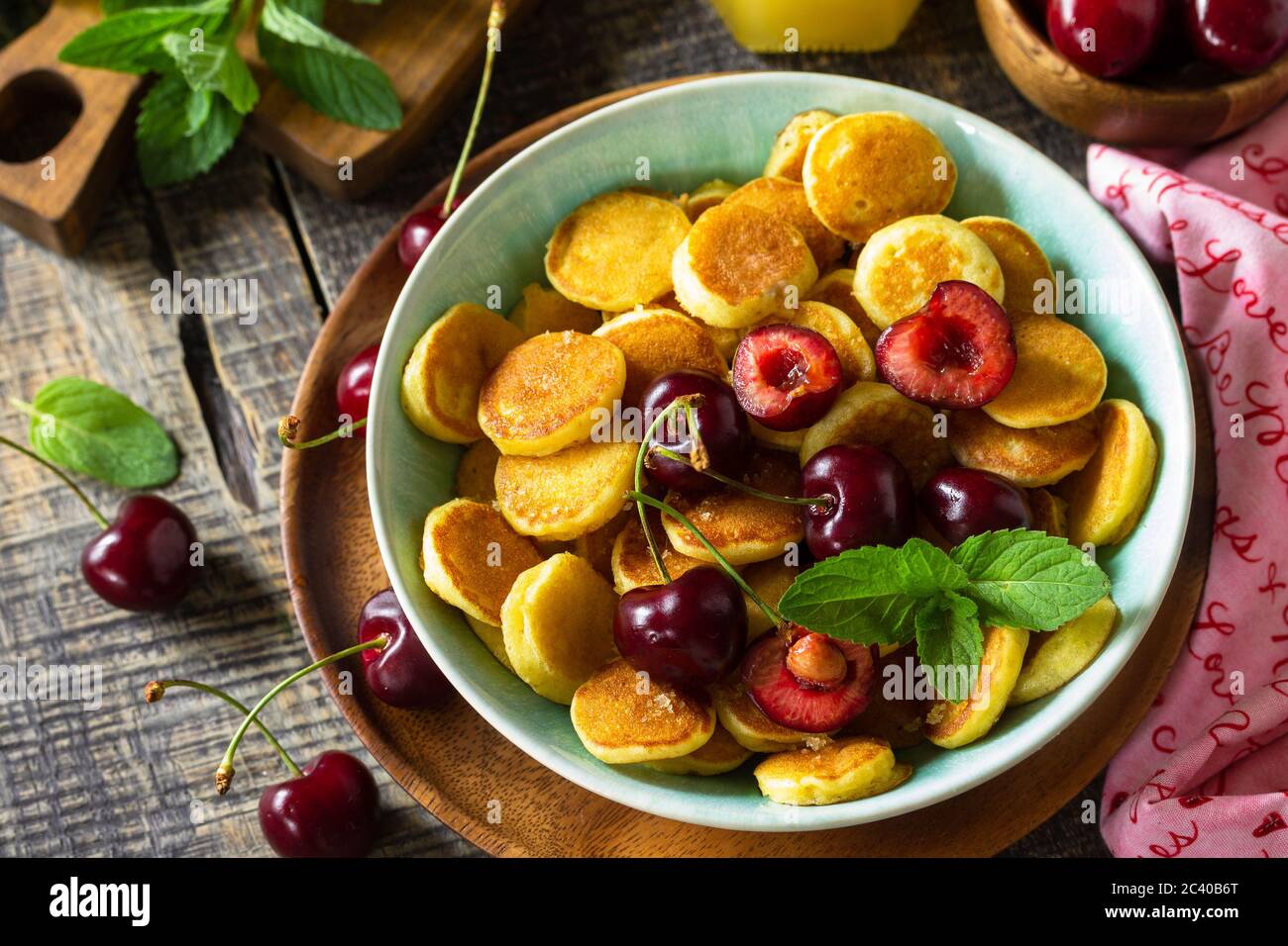 Minuscoli pancake, cibo alla moda. Mini pancake con ciliegie e miele su un rustico tavolo in legno. Foto Stock