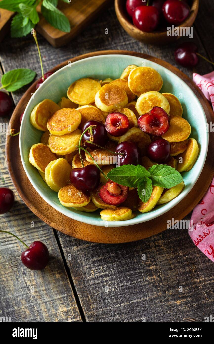 Minuscoli pancake, cibo alla moda. Mini pancake con ciliegie e miele su un rustico tavolo in legno. Spazio di copia. Foto Stock