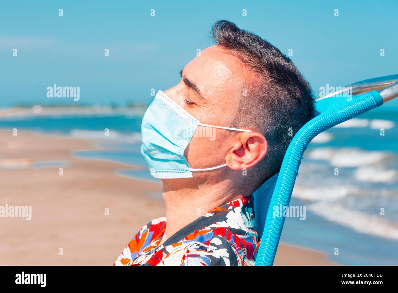 primo piano di un uomo caucasico che indossa una maschera chirurgica blu mentre è seduto su una sedia a sdraio sulla spiaggia, con il mare sullo sfondo Foto Stock