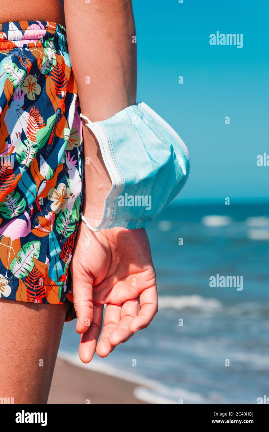 primo piano di un uomo caucasico, visto da dietro, in costume da bagno in piedi sulla spiaggia, indossando una maschera chirurgica blu nel braccio, come sta prendendo un sfiatatoio di w. Foto Stock