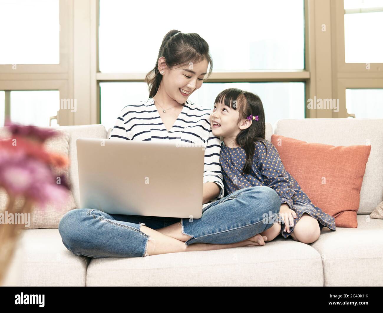 giovane donna asiatica d'affari che lavora da casa seduto sul divano usando il computer portatile mentre si prende cura della figlia Foto Stock