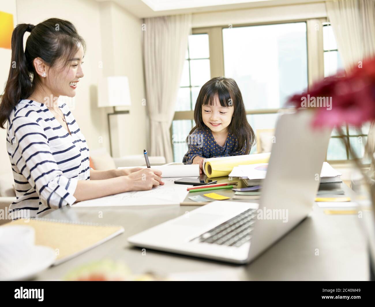 giovane designer asiatico madre che lavora da casa mentre si prende cura di figlia (grafica in background alterata digitalmente) Foto Stock