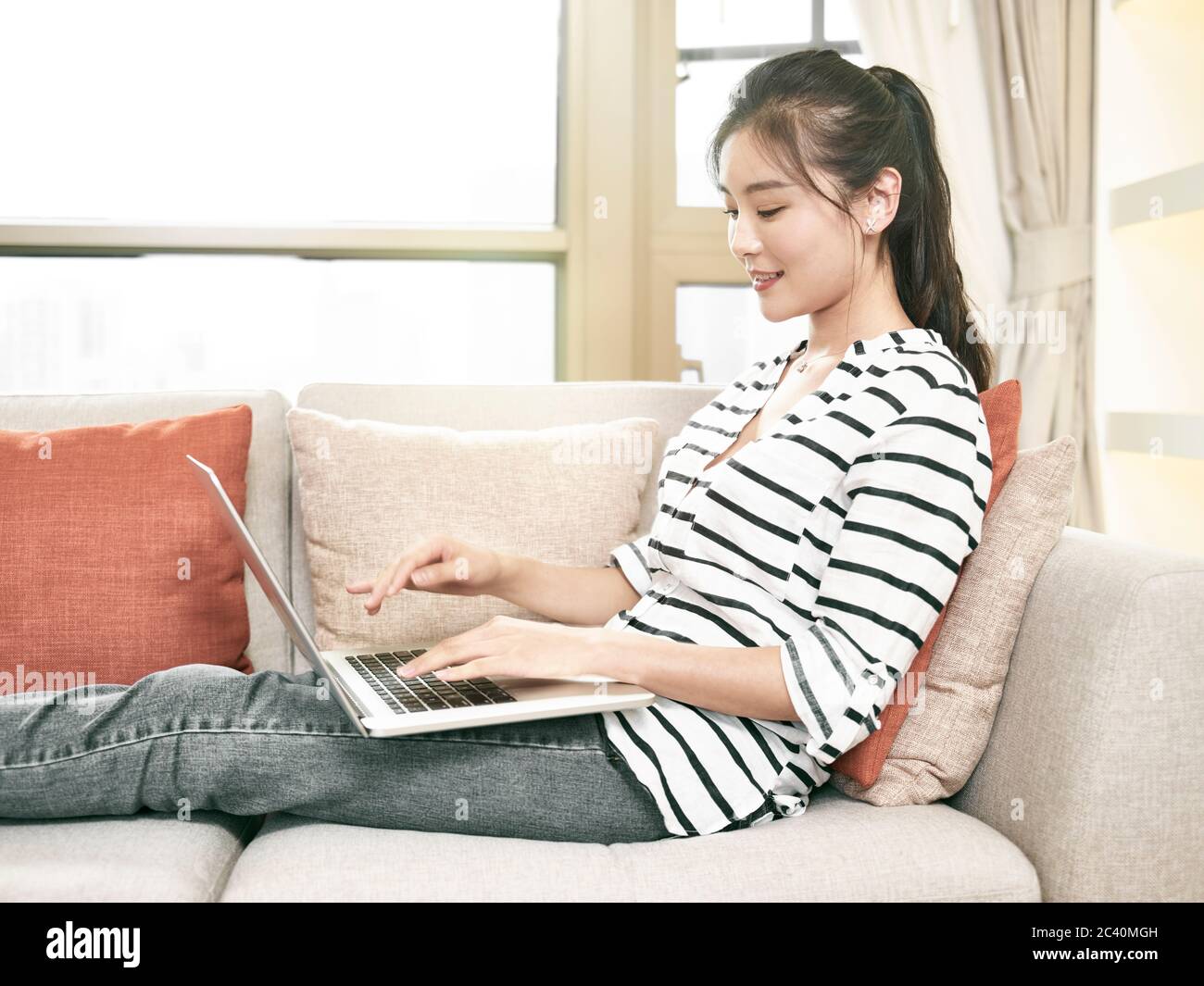 giovane donna asiatica che lavora da casa seduta sul divano usando un computer portatile Foto Stock