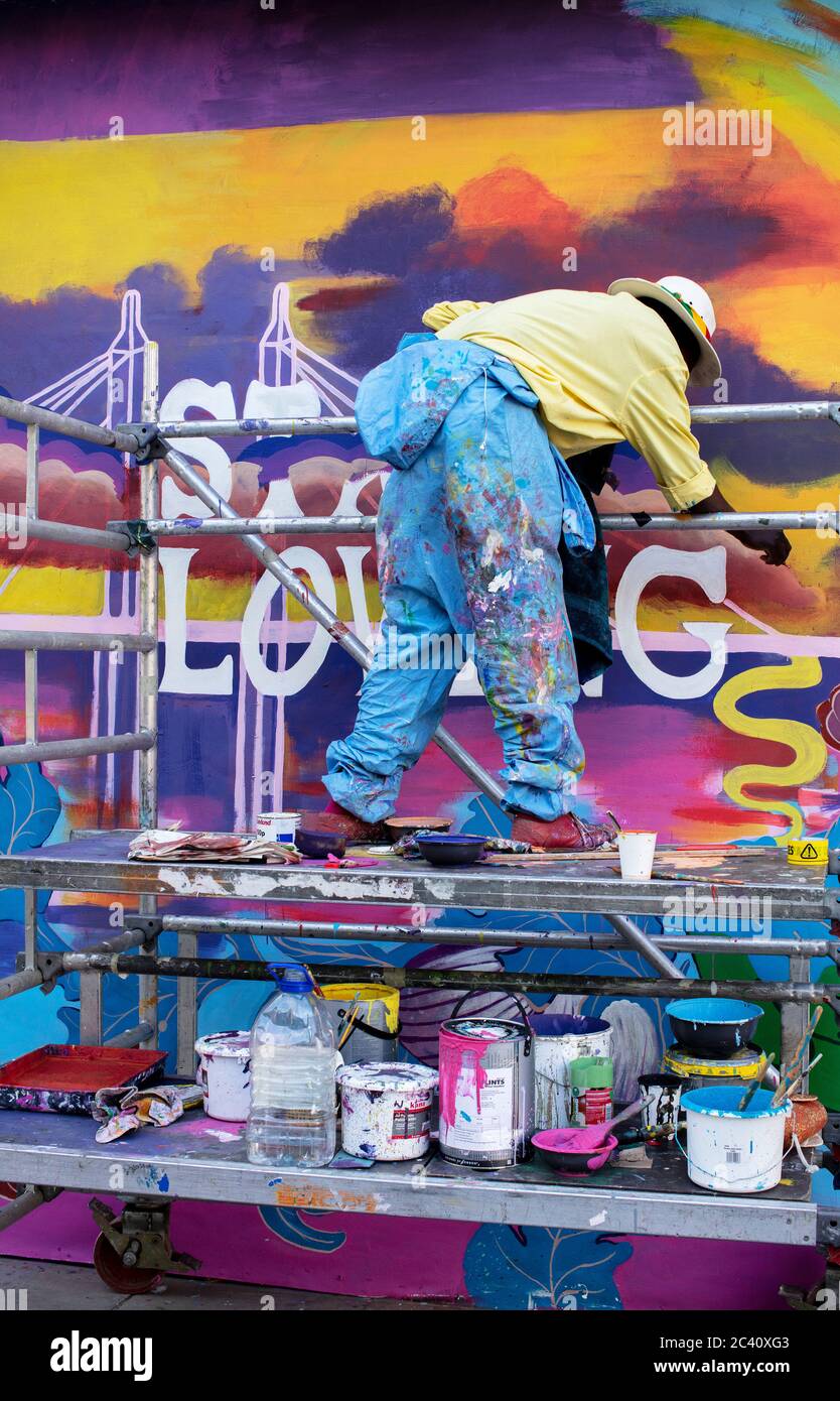 Uomo su piattaforma impalcata dipingendo un'accaparramento su King's Road, Chelsea, Londra. Tini di vernice multicolore e pantaloni e scarpe lacernati Foto Stock