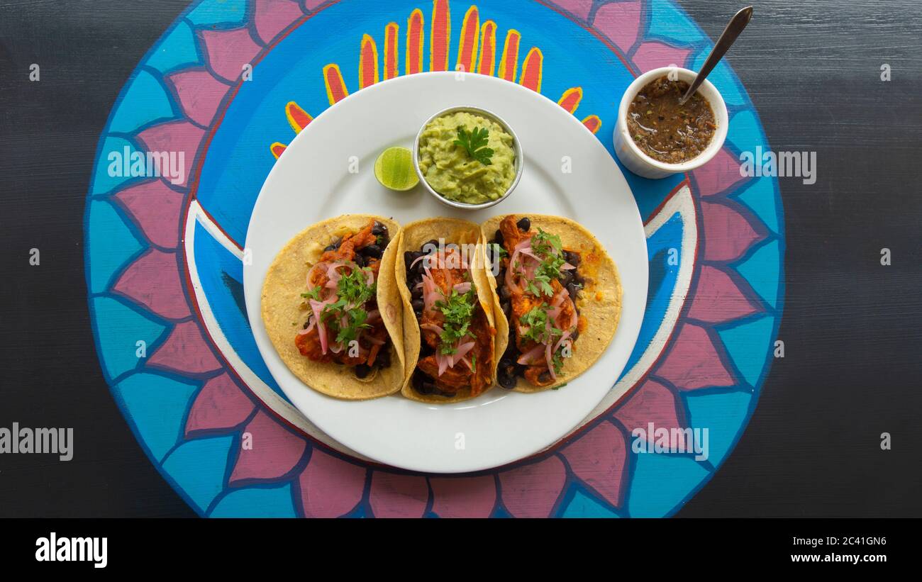 Vista dall'alto di un piatto bianco con tre tacos messicani di carne accompagnati da una piccola pentola di guacamole e salsa piccante su un tavolo di legno con vari colori Foto Stock