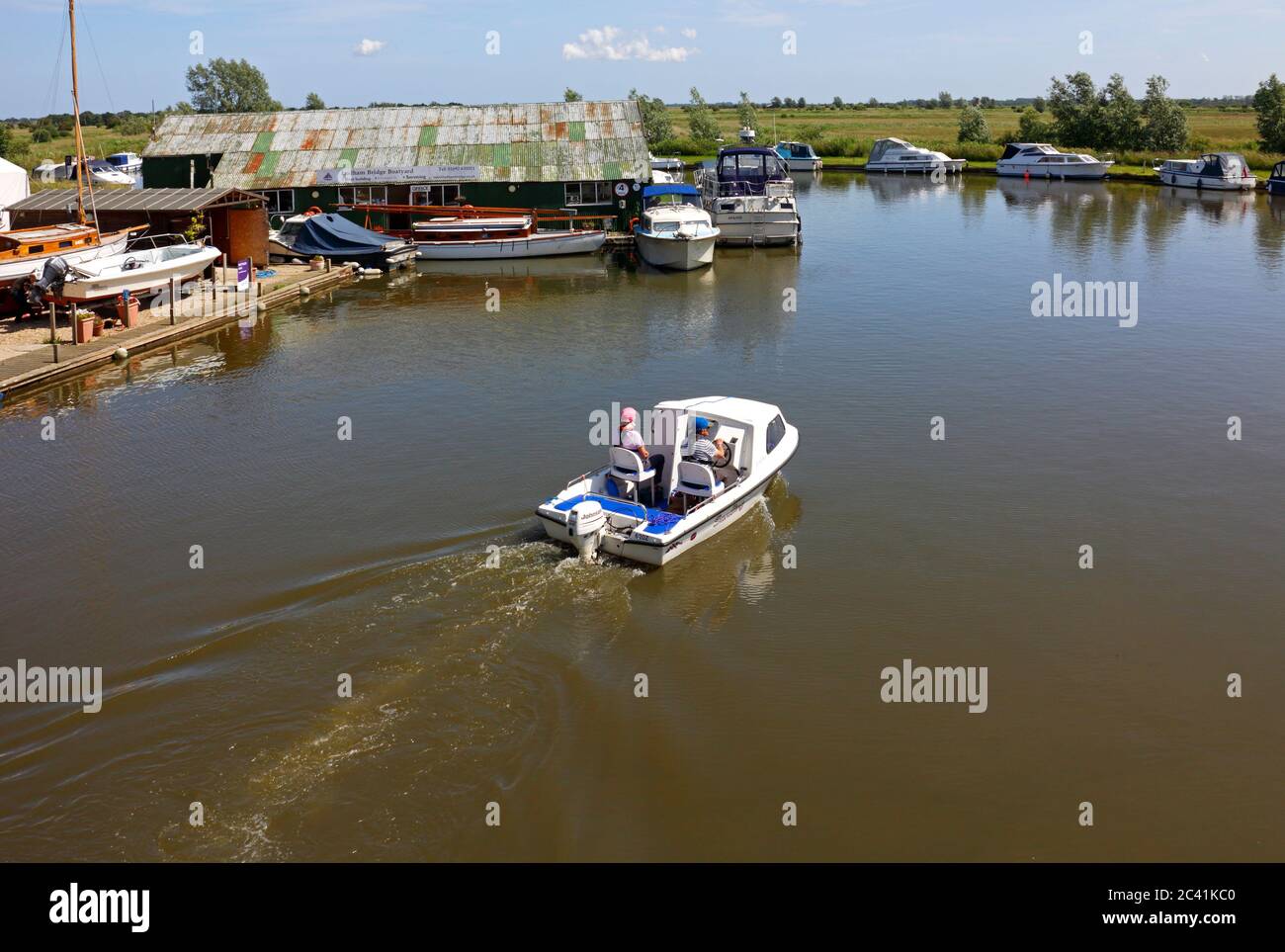 Una coppia in un piccolo mestiere da diporto che passa davanti ad un cantiere sul fiume ANT sulla Norfolk Broads a Ludham, Norfolk, Inghilterra, Regno Unito. Foto Stock