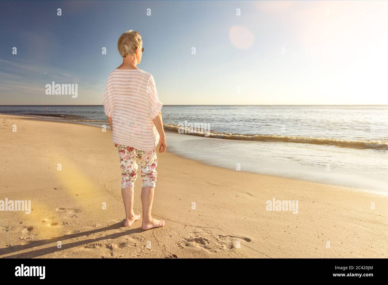 Donna anziana in piedi sulla spiaggia e guardando verso l'oceano con un'atmosfera di lente scenografica Foto Stock