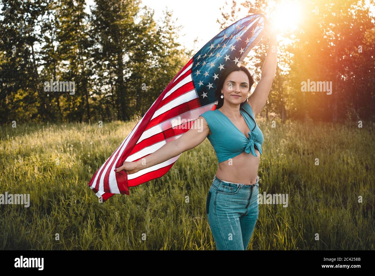 Donna che indossa jeans e la parte superiore blu che tiene una bandiera americana sopra la sua testa su una natura durante la celebrazione del tramonto. Foto Stock