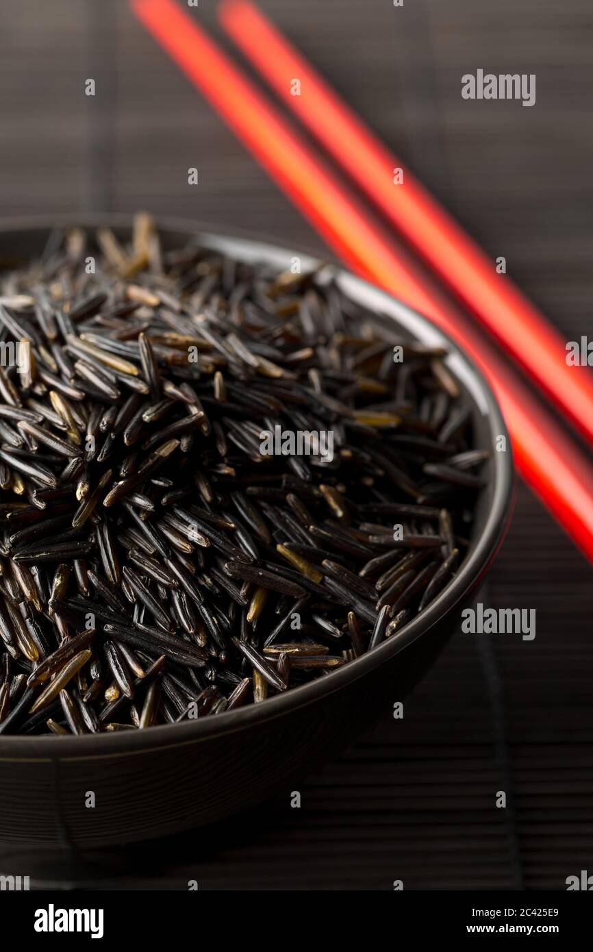 Mucchio di grani di riso selvatico, crudo e nero non cotti in ciotola nera con i chopsticks rossi sullo sfondo nero del tappeto di bambù, fuoco selettivo Foto Stock