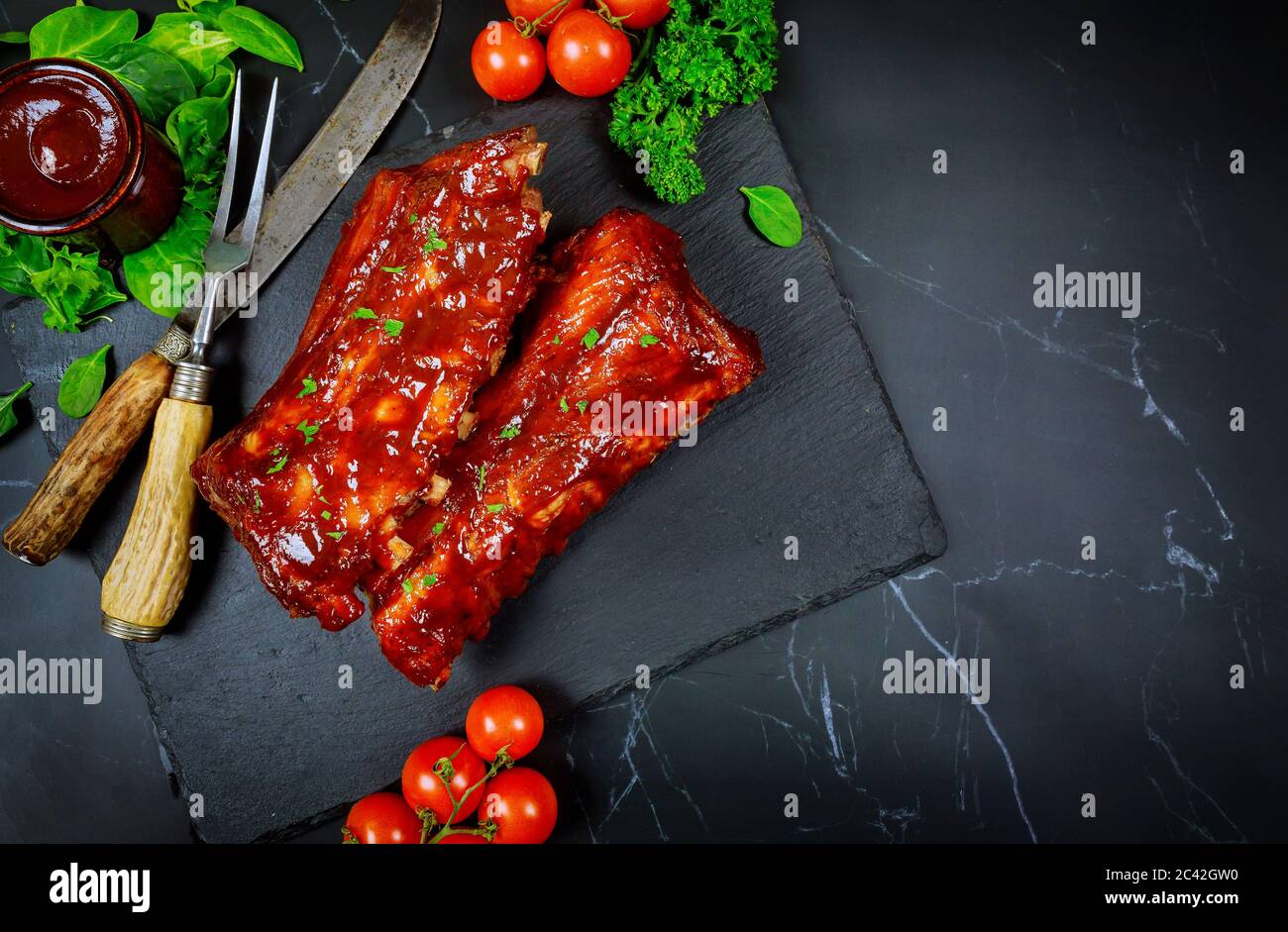 Costolette di maiale grigliate con salsa barbecue, pomodoro e spinaci su tavola nera. Foto Stock