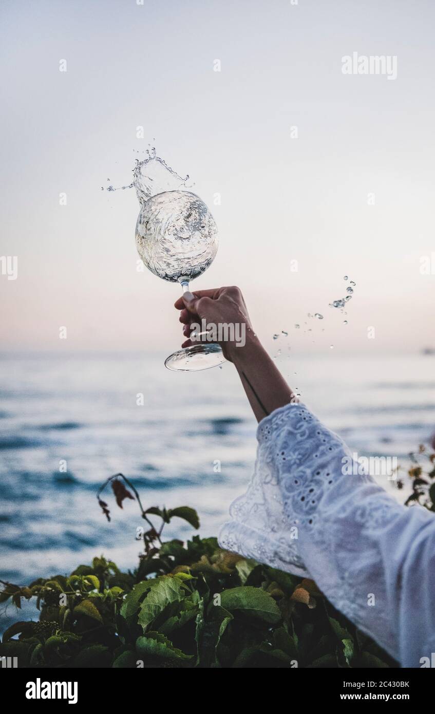 Mano femminile in pizzo bianco manica canica girando bicchiere di vino bianco scorrevole con gocce e bei colori del tramonto e orizzonte di mare sullo sfondo. Summe Foto Stock