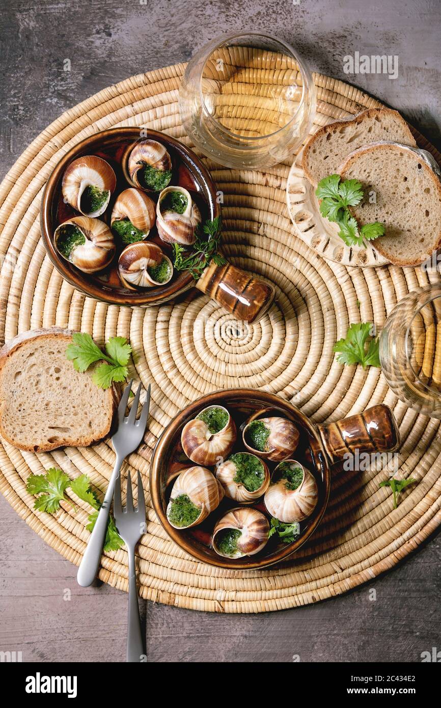 Escargots de Bourgogne - lumache con burro alle erbe, piatto gourmet, in tradizionali padelle di ceramica con coriandolo, pane, bicchiere di vino bianco su napki paglia Foto Stock