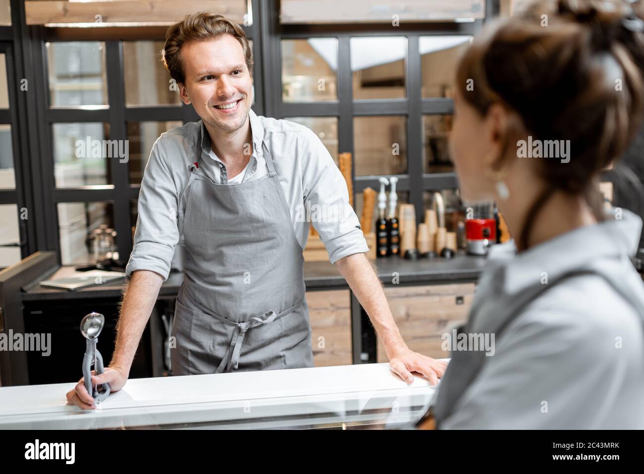 Ritratto di un allegro venditore in grembiule che parla con un cliente al piccolo negozio o al bar. Concetto di piccola impresa e lavoro nel campo dei servizi Foto Stock