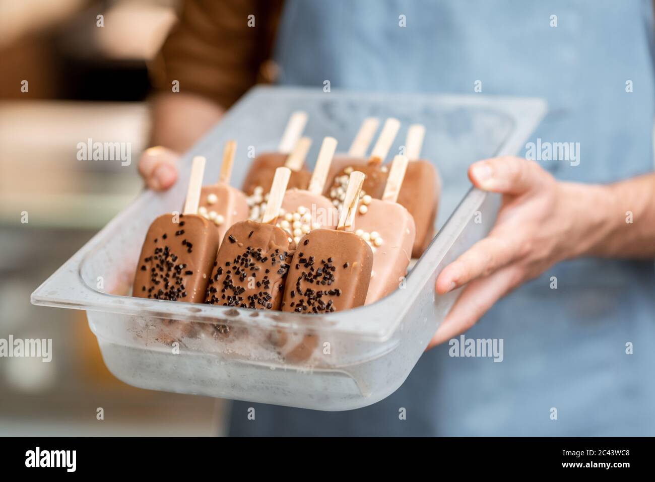 Venditore che tiene un vassoio con gelato al cioccolato su un bastone nel negozio, primo piano su gelato su uno sfondo blu grembiule Foto Stock