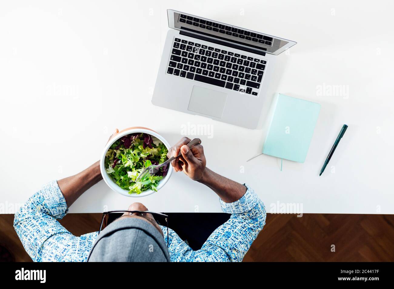 Giovane uomo che mangia insalata mentre si siede con il computer portatile alla scrivania dell'ufficio di casa Foto Stock