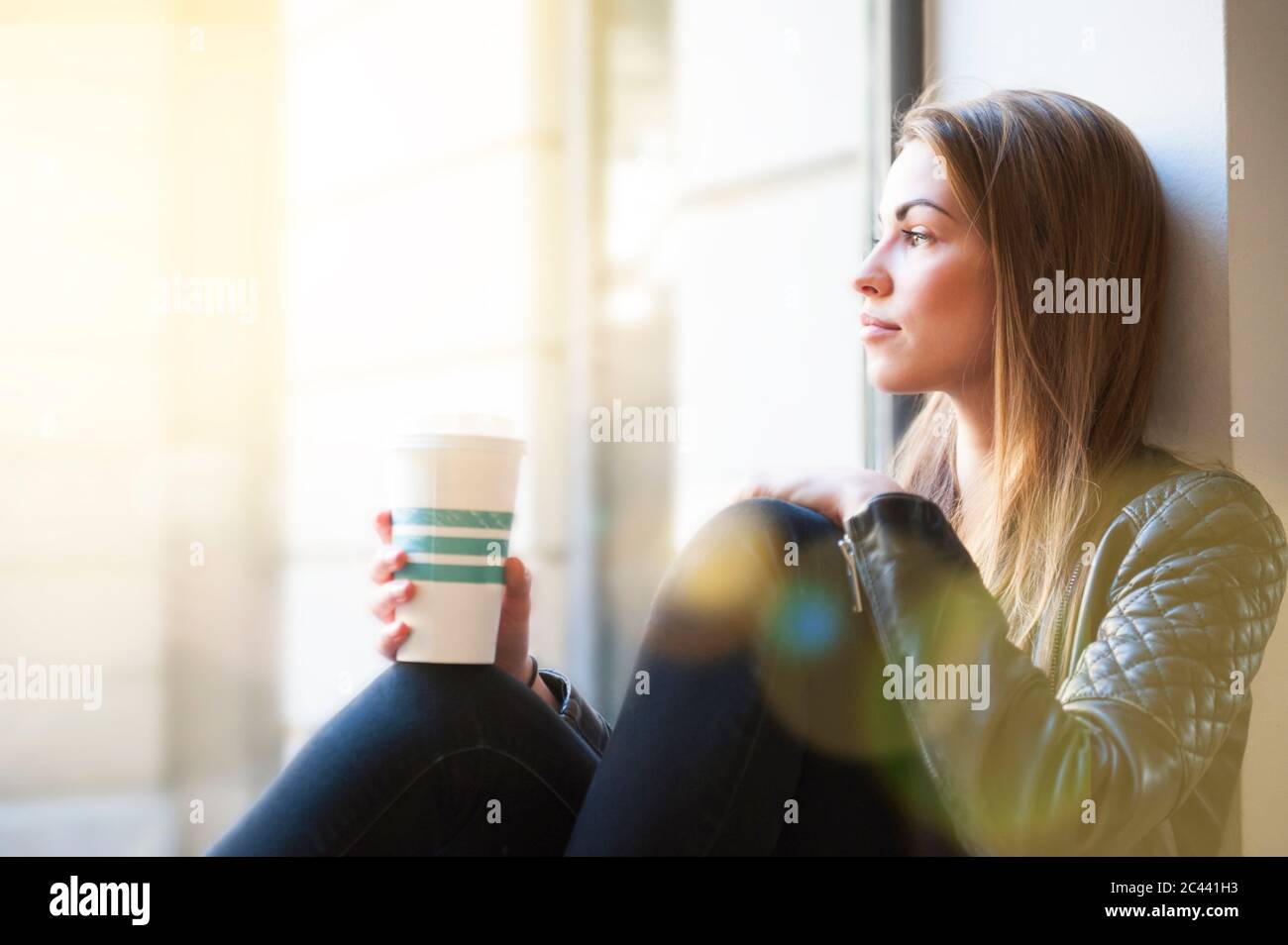 Donna che guarda attraverso la finestra mentre tiene una tazza di caffè usa e getta nel bar Foto Stock