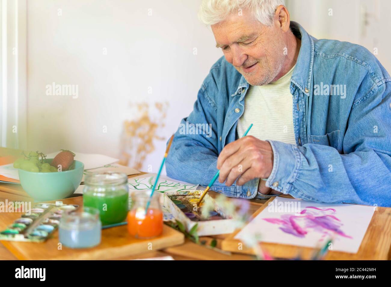Felice uomo anziano pittura con vernici acquerello sul tavolo a casa Foto Stock