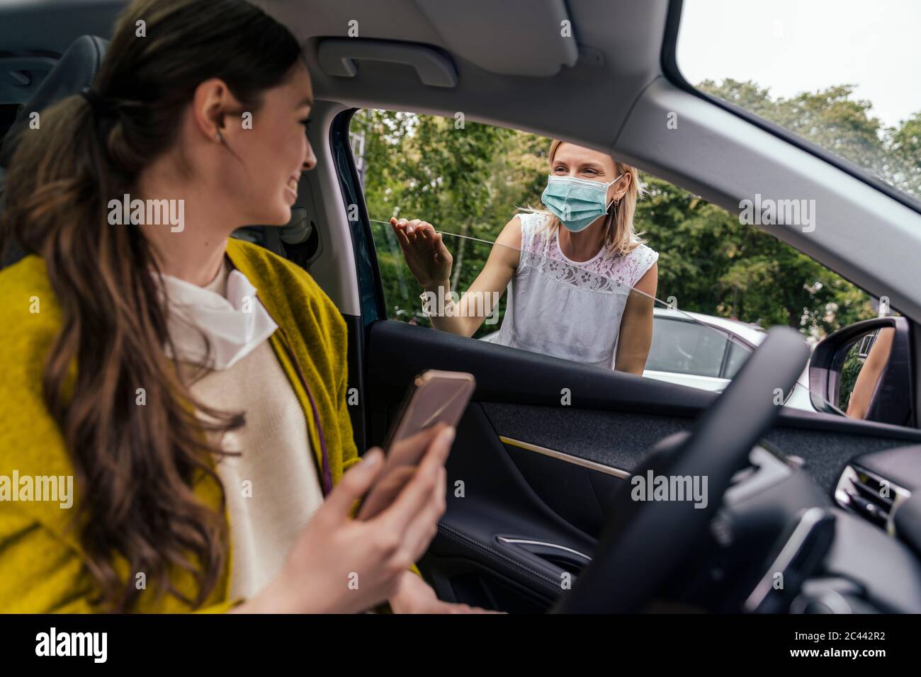 Donna che indossa una maschera protettiva che parla attraverso la finestra dell'auto con una donna senza maschera Foto Stock