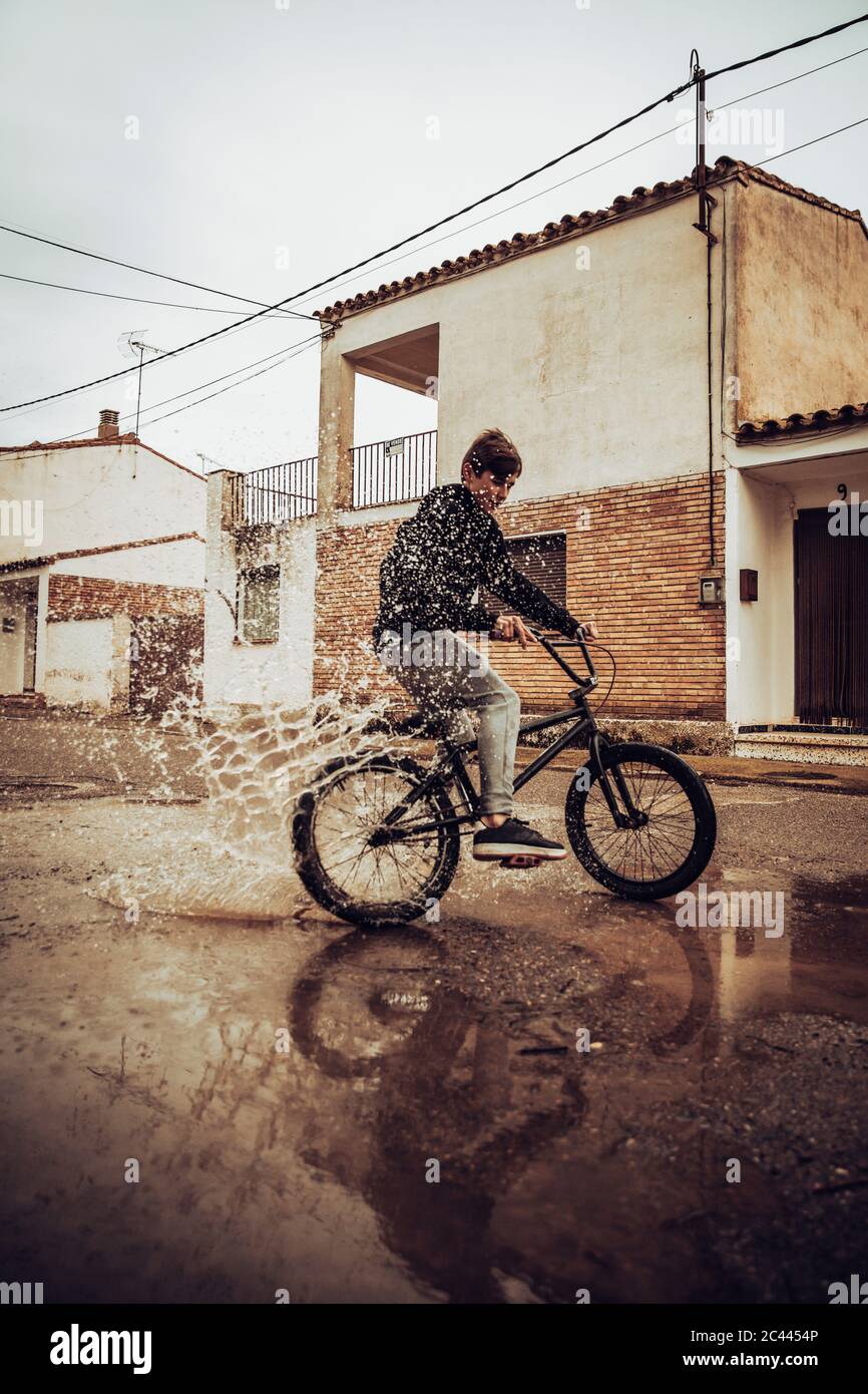 Ragazzo adolescente che spruzzi acqua in puddle mentre cavalcano in bicicletta sulla strada durante la stagione delle piogge Foto Stock