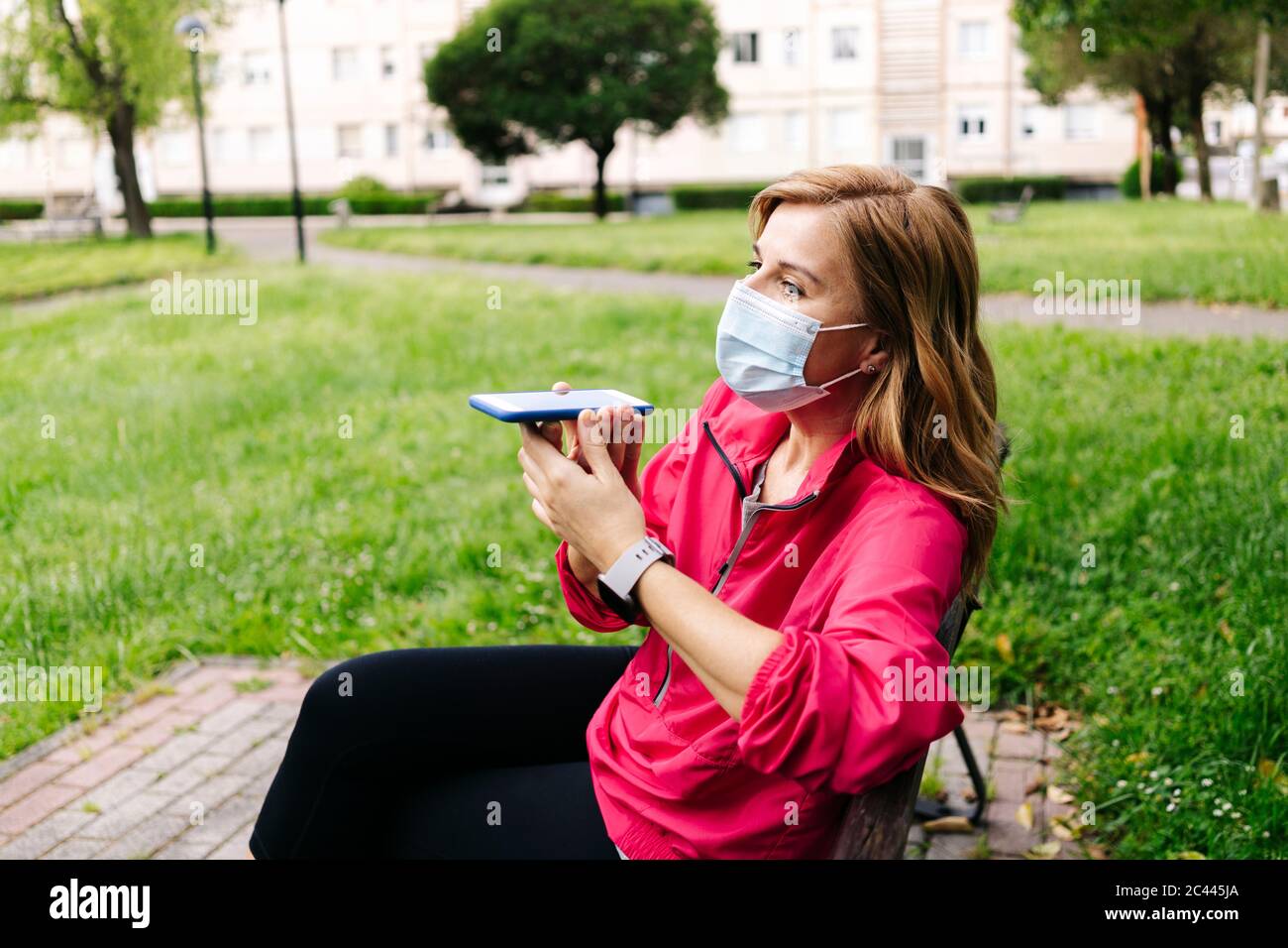 Donna con maschera chirurgica con telefono a mani libere seduto sul banco Foto Stock