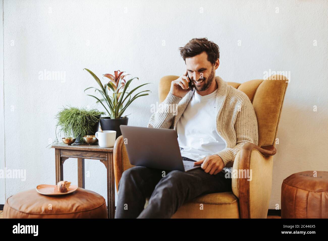 Uomo sorridente sul telefono seduto sulla poltrona a casa guardando il computer portatile Foto Stock