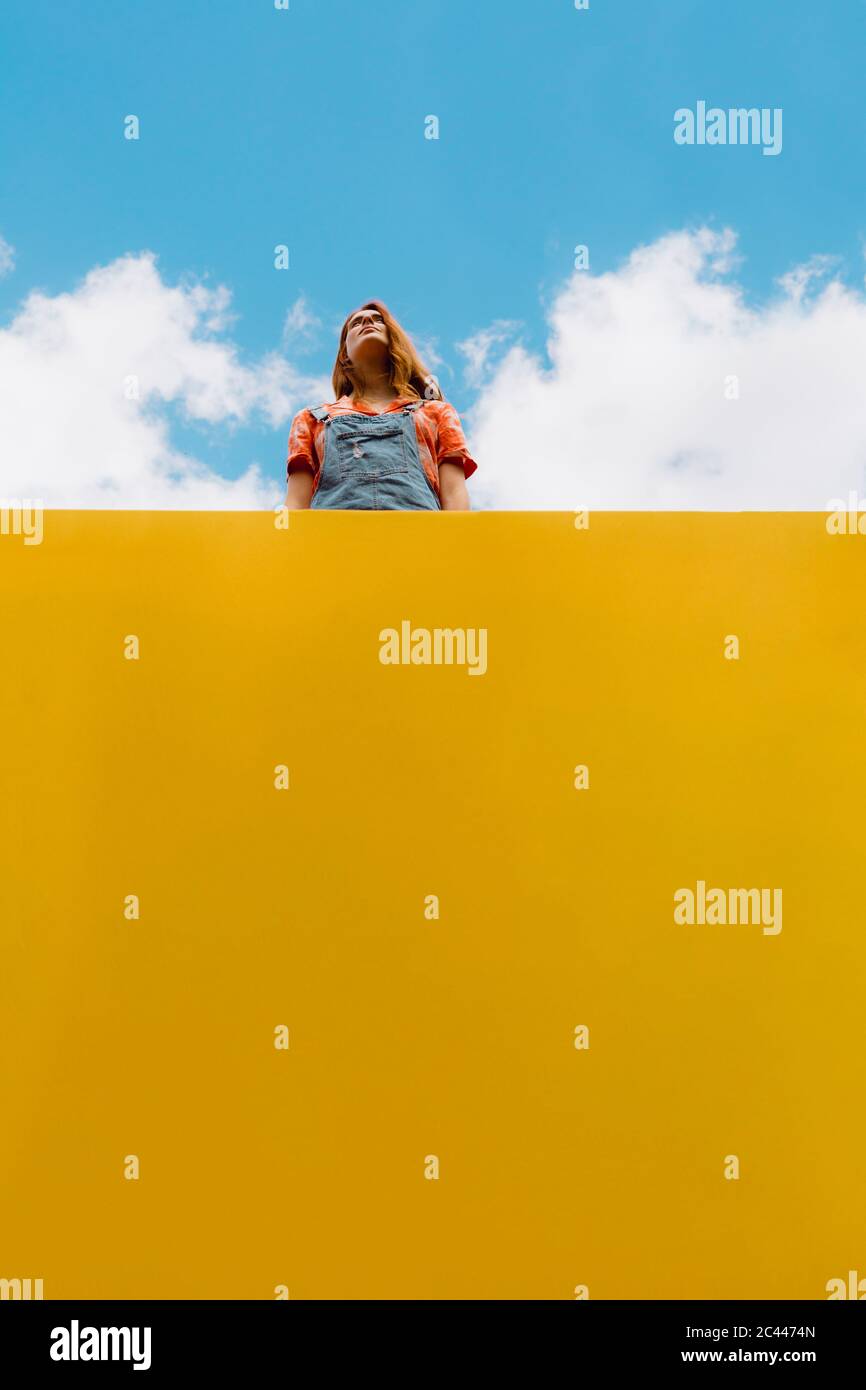 Giovane donna in piedi sopra la parete gialla contro il cielo nuvoloso Foto Stock