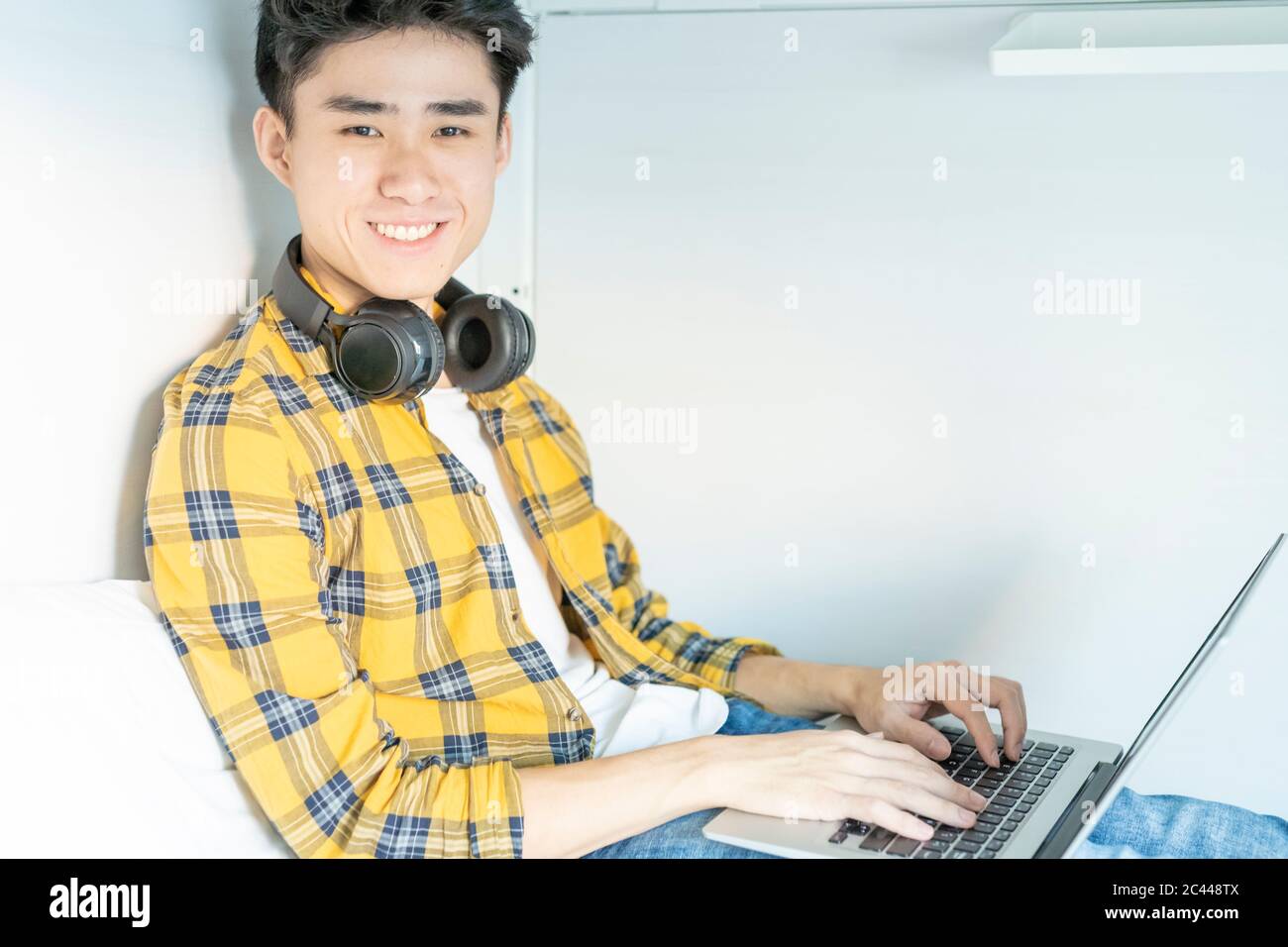 Ritratto di un giovane sorridente con le cuffie seduti a letto utilizzando un computer portatile Foto Stock