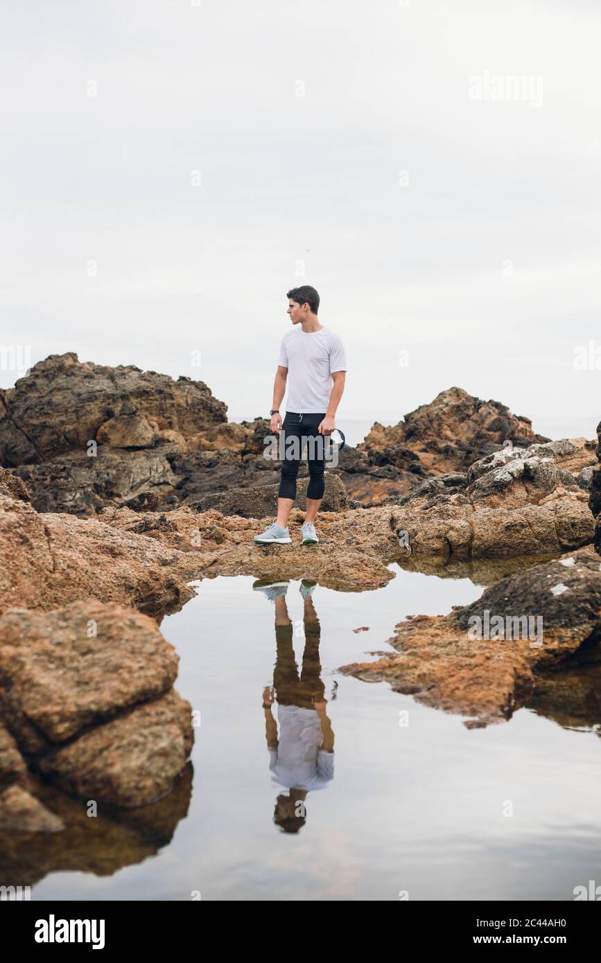 Lunghezza completa di corridore di sentiero maschile in piedi su rocce con il suo riflesso sull'acqua contro il cielo, Ferrol, Galizia, Spagna Foto Stock