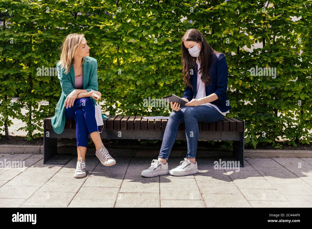 Due uomini d'affari seduti sulla panchina all'esterno e mantenendo la distanza mentre indossano la maschera facciale Foto Stock
