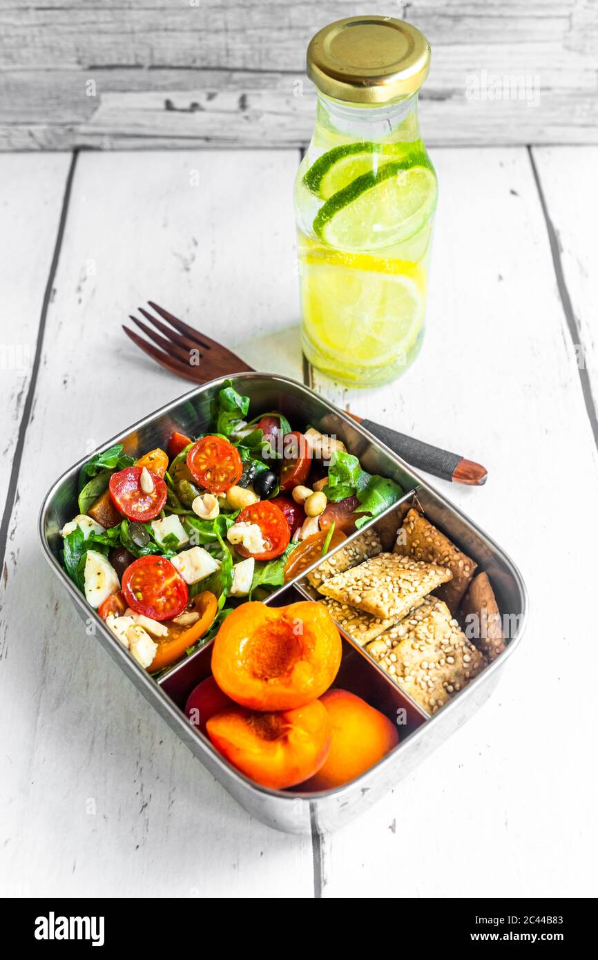 Pranzo al sacco con insalata di razzi con pomodori colorati, mozzarella e noci, croccante e albicocche e bottiglia d'acqua infusa con limoni e fette di lime Foto Stock