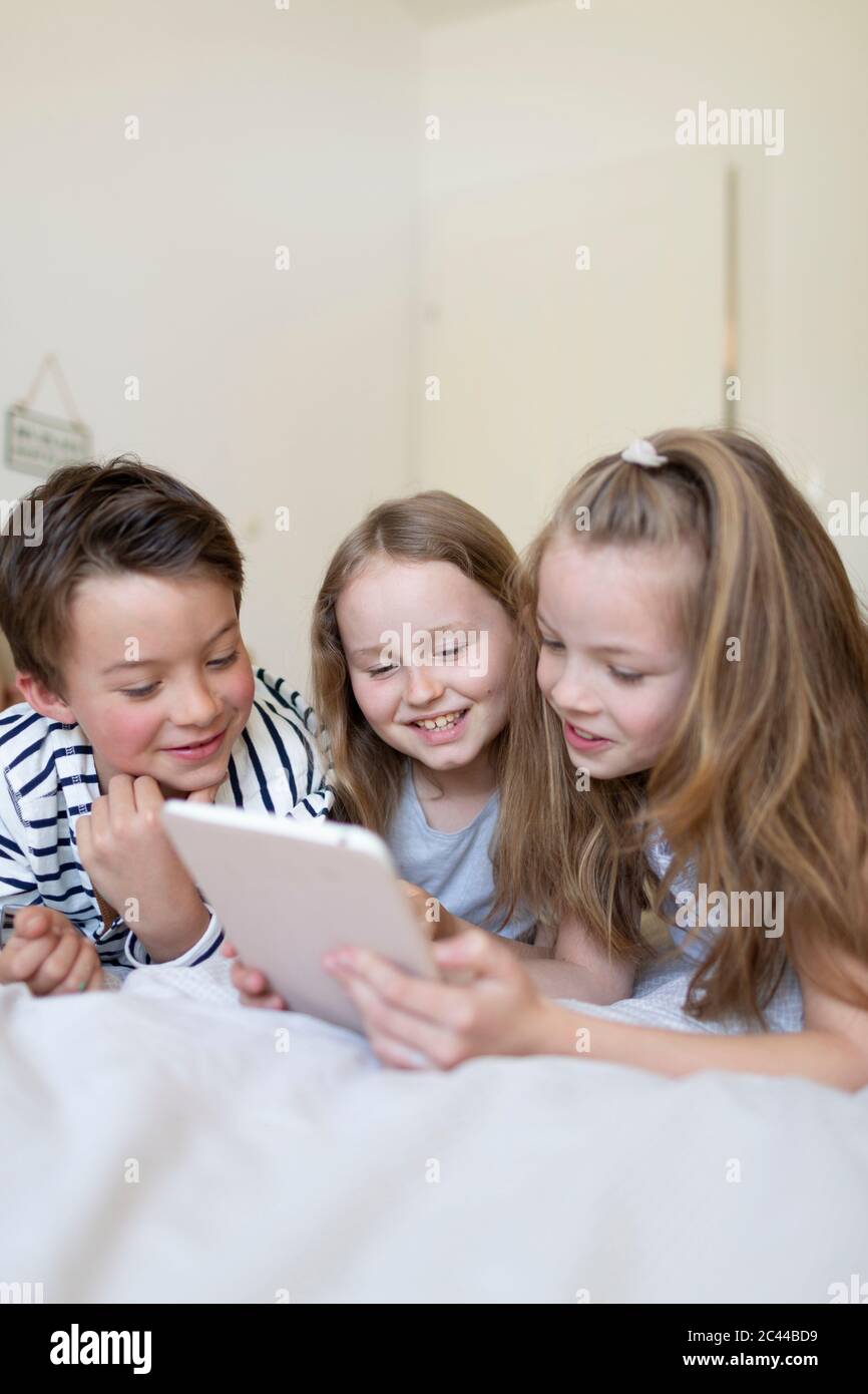 Ritratto di ragazzo e delle sue due sorelle che si stengono a letto usando un tablet digitale per la casa Foto Stock