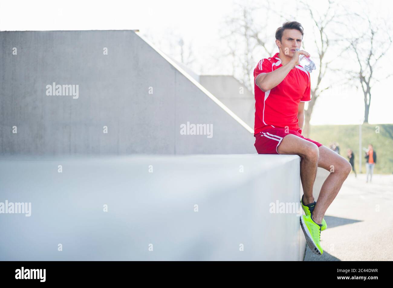 Uomo medio adulto che beve acqua guardando via mentre si siede su muro di ritegno Foto Stock