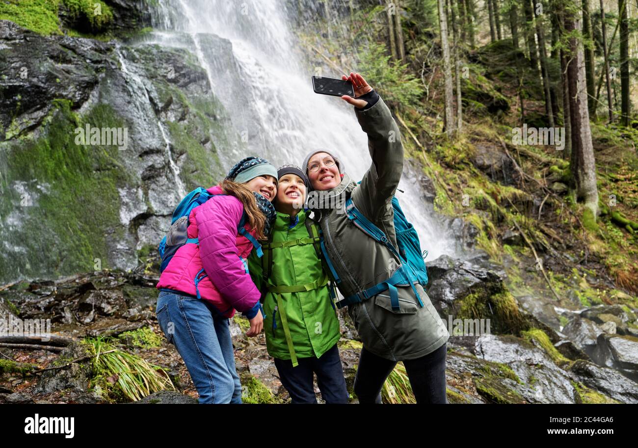 Donna sorridente che prende selfie con bambini contro la cascata Zweribach nella Foresta Nera settentrionale Foto Stock