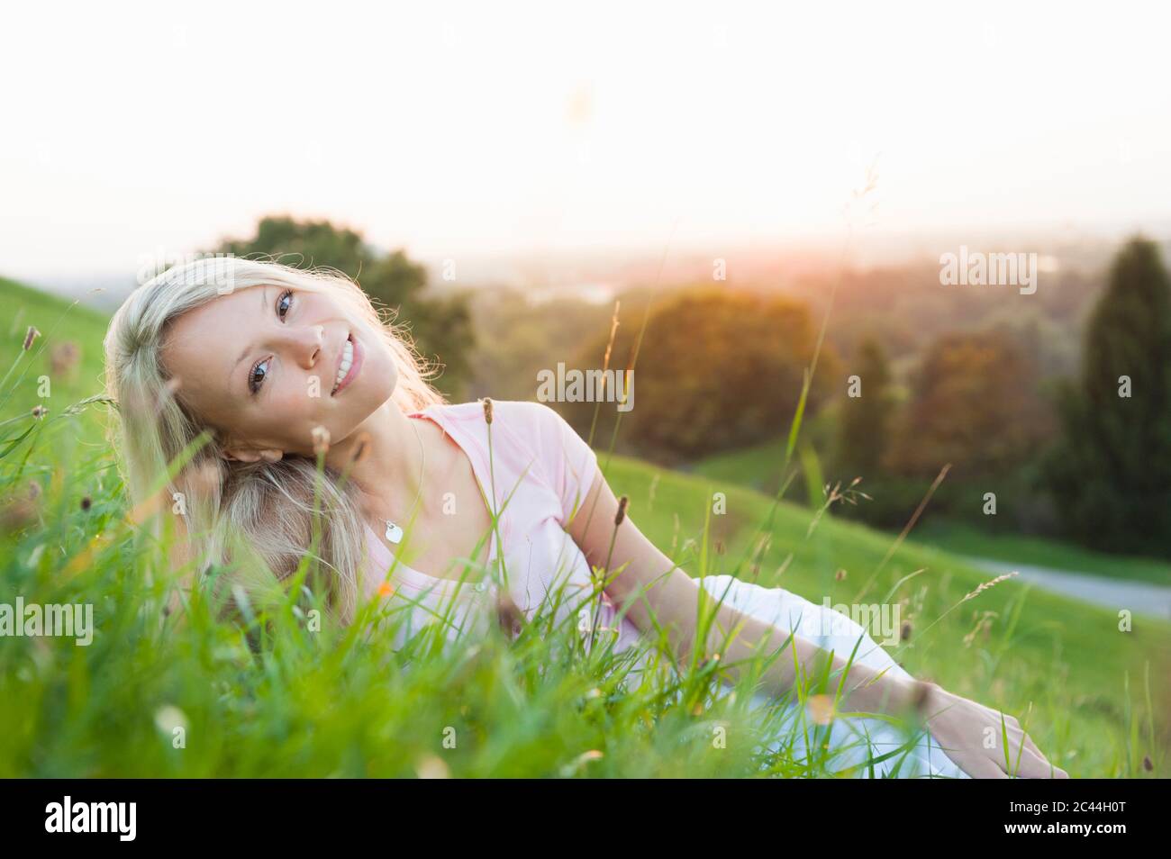 Sorridente giovane bella donna che si stende su terra erbosa contro il cielo limpido durante il tramonto Foto Stock
