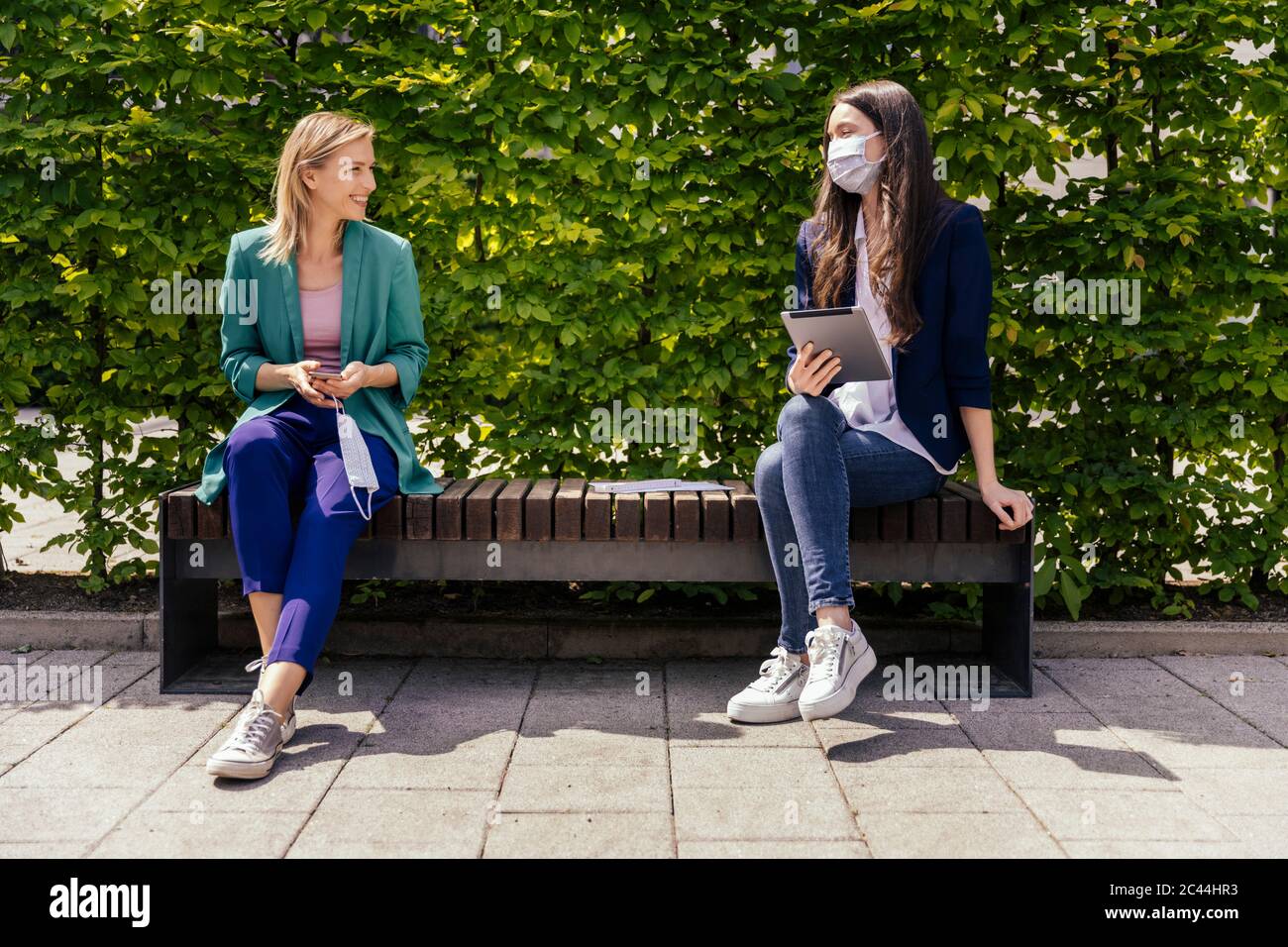 Due uomini d'affari seduti sulla panchina all'esterno e mantenendo la distanza mentre indossano la maschera facciale Foto Stock