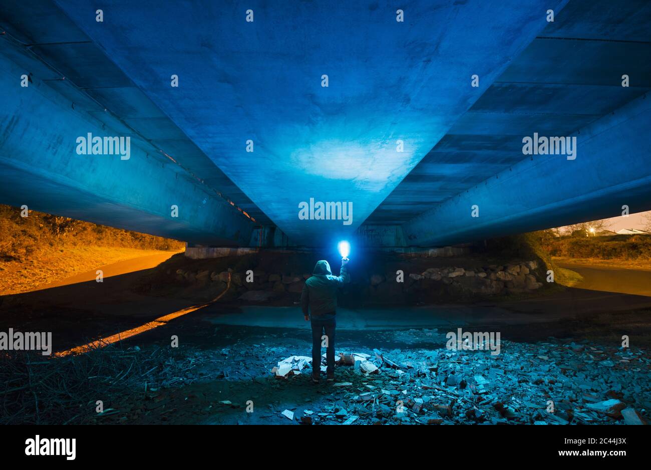 Spagna, Galizia, Vista posteriore di un uomo con cappuccio sotto un ponte di cemento con una luce blu brillante in mano Foto Stock