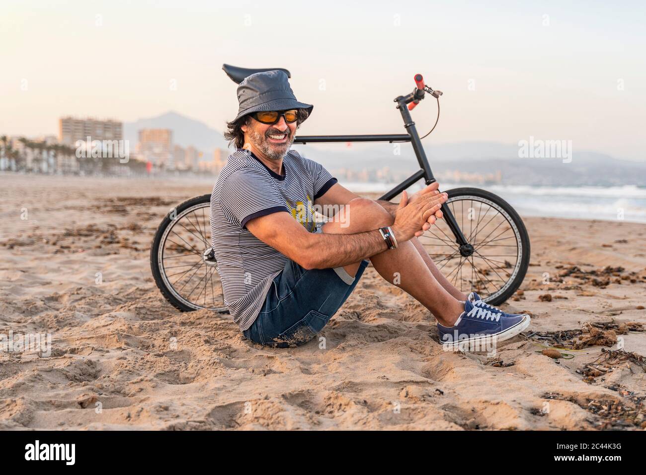 Uomo maturo con bicicletta, seduto sulla spiaggia, sorridente Foto Stock