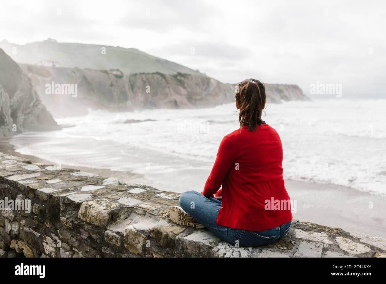 Vista posteriore completa di turisti rilassati seduti su un muro di contenimento mentre si guarda le scogliere contro il cielo, Itzurun, Zumaia, Spagna Paesi Baschi Foto Stock