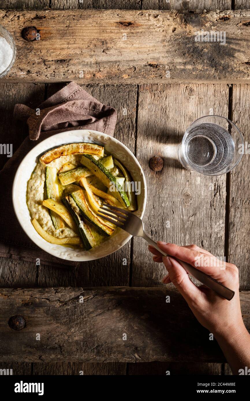 Ritagliate la mano della donna mangiare hummus con zucchine fritte sul tavolo di legno Foto Stock