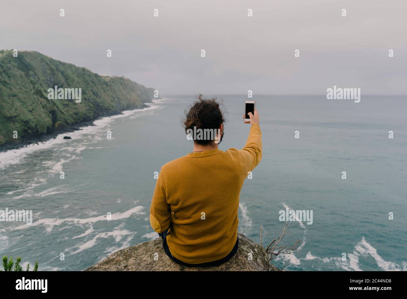 Vista posteriore di un uomo seduto su una roccia sulla costa scattando una foto su un smartphone sull'isola di Sao Miguel, Azzorre, Portogallo Foto Stock