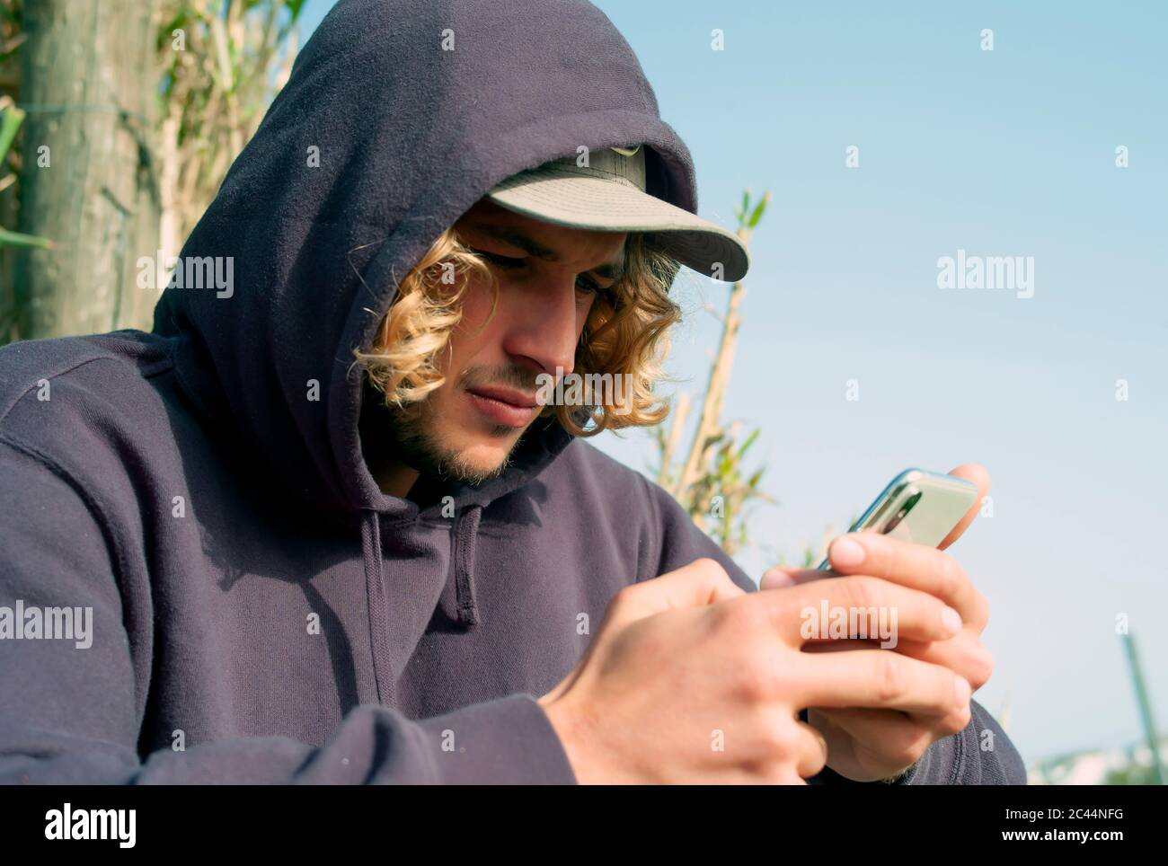 Primo piano di un giovane uomo che indossa il cappuccio utilizzando uno smartphone mentre si rilassa alla spiaggia Tarifa, Spagna Foto Stock