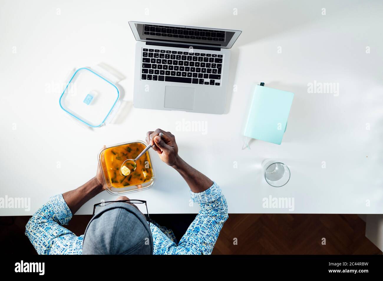 Giovane uomo che mangia la zuppa dal contenitore mentre si siede con il computer portatile alla scrivania dell'ufficio di casa Foto Stock