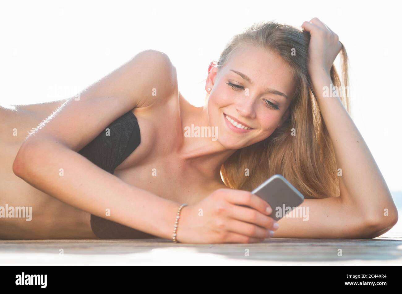 Ritratto di giovane donna sorridente sdraiata sul molo guardando il telefono cellulare Foto Stock