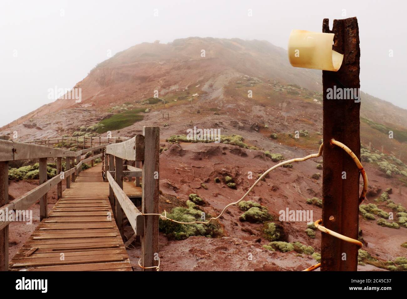 Palo arrugginito con etichetta gialla su un passerella di legno che conduce ad una montagna nebbiosa Foto Stock