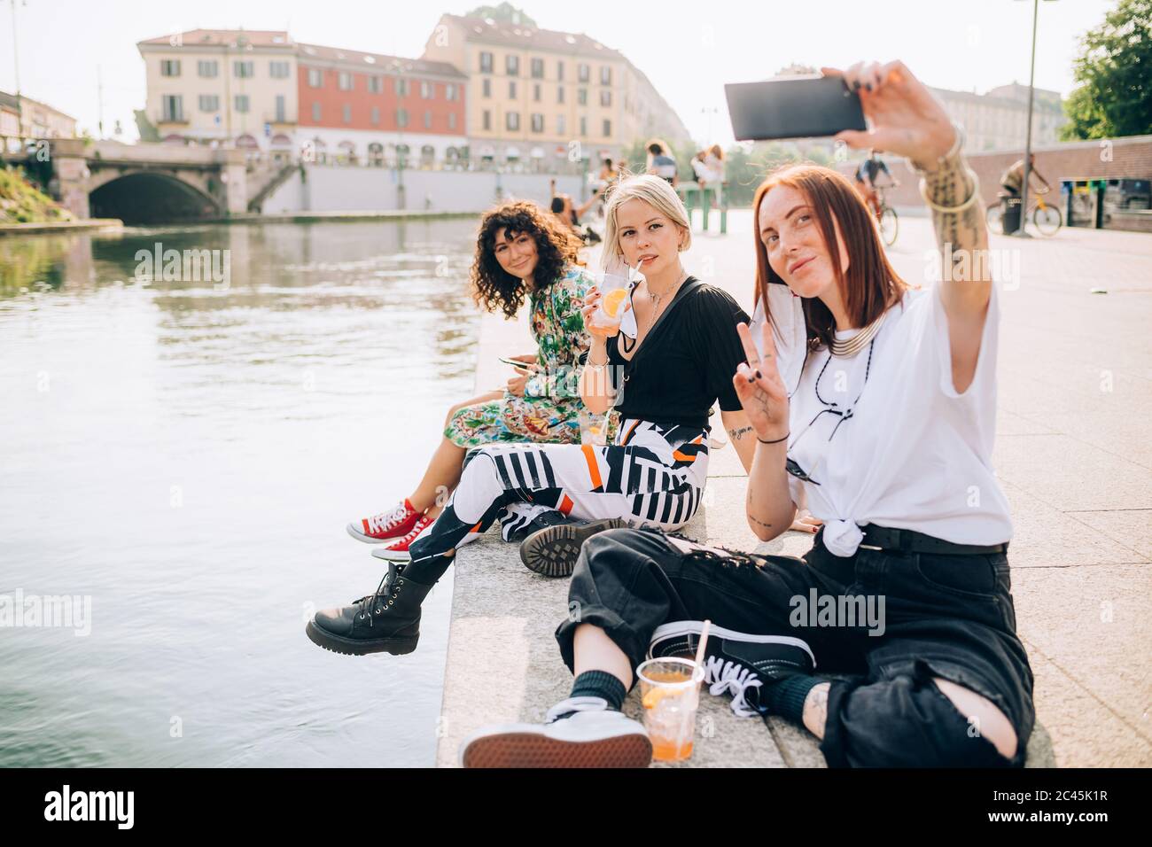 Tre giovani donne sedute su una riva del fiume, che prendono selfie. Foto Stock