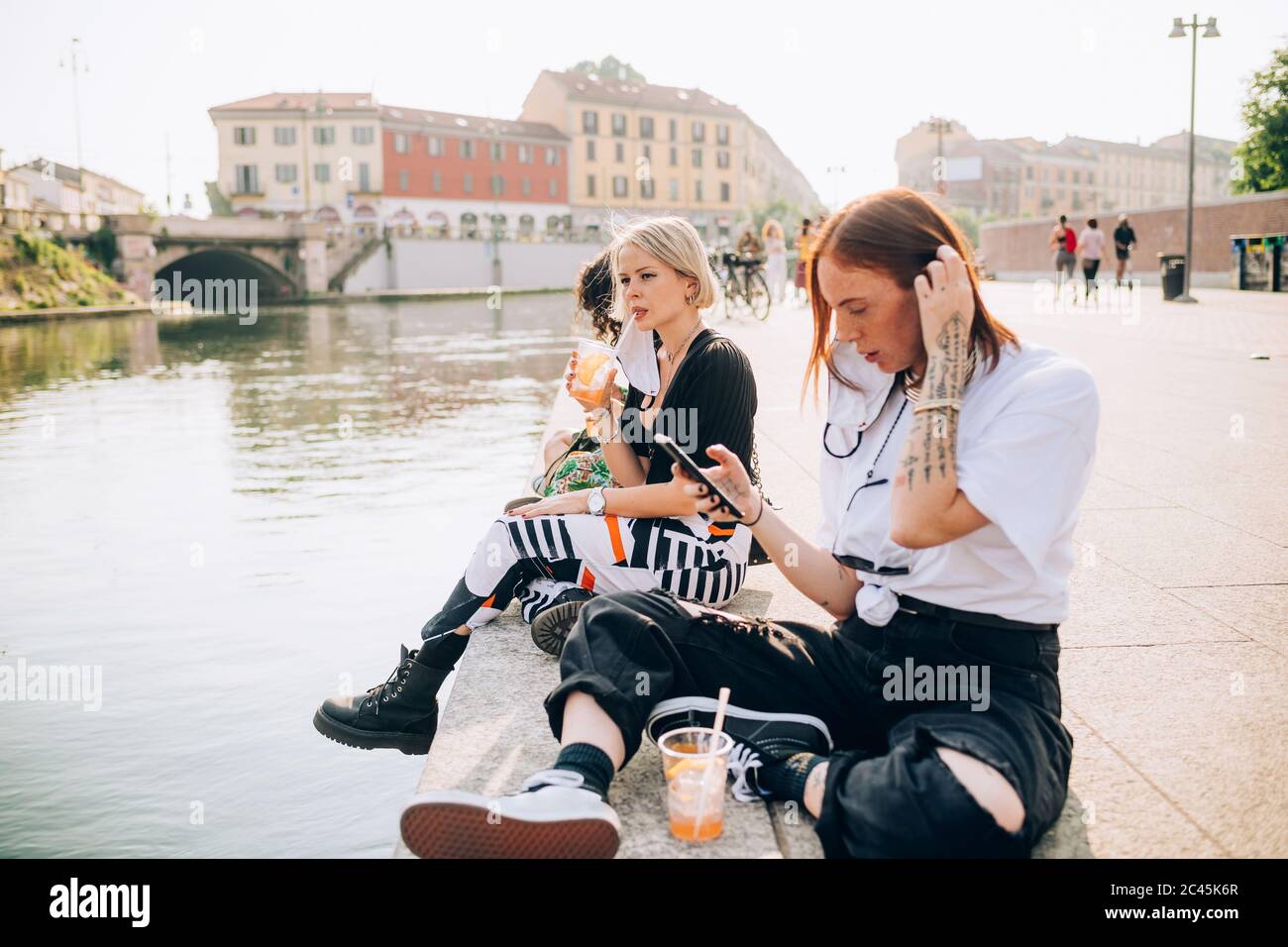 Tre giovani donne sedute su una riva del fiume, bevendo e utilizzando il telefono cellulare. Foto Stock