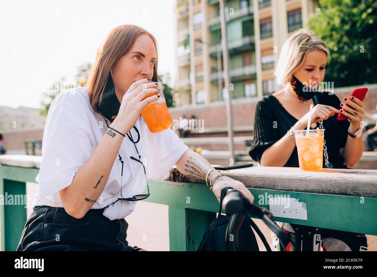 Due giovani donne sedute all'aperto su una panca, bevendo e utilizzando il telefono cellulare. Foto Stock
