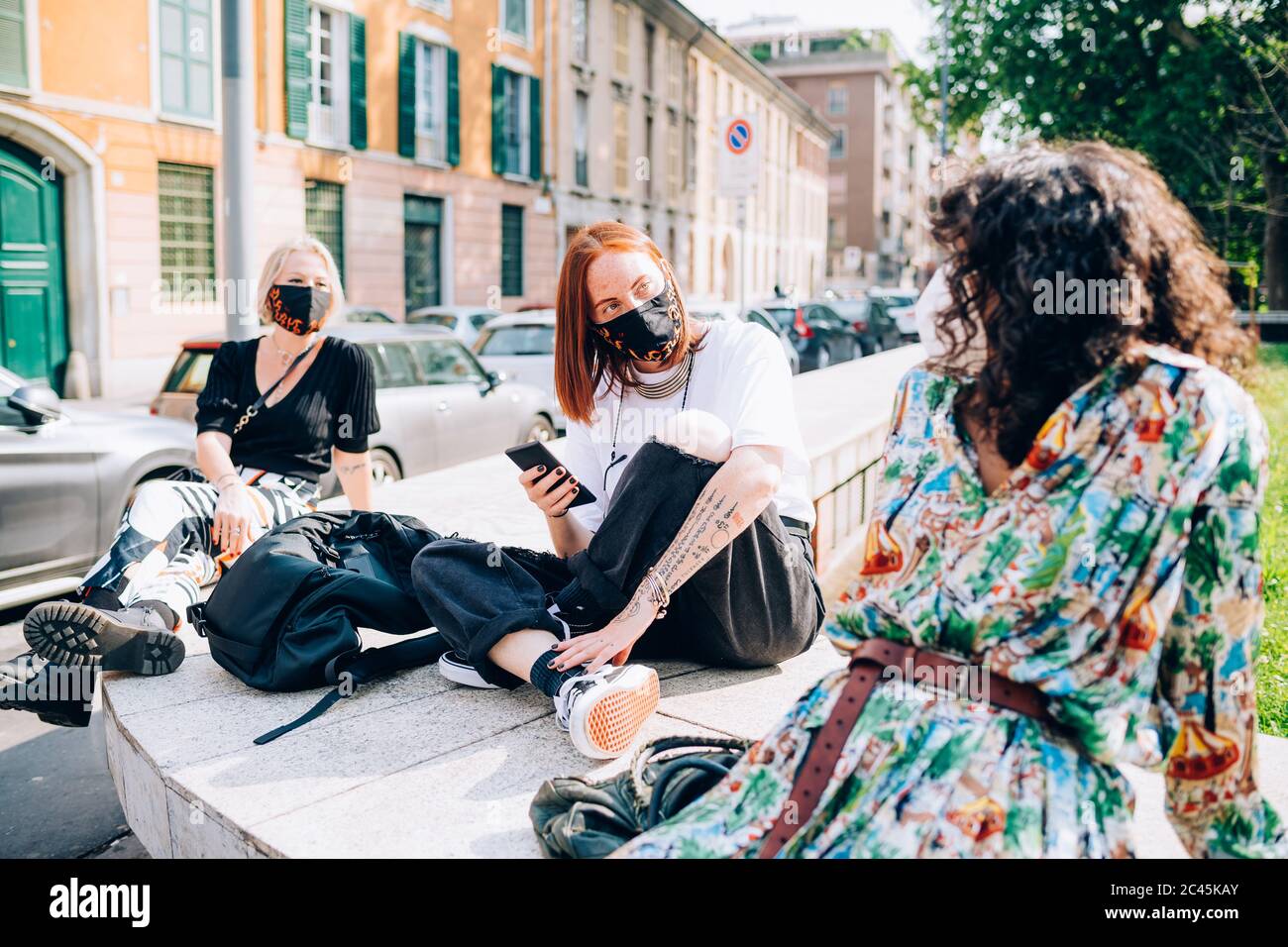Tre giovani donne che indossano maschere di fronte durante il virus Corona, seduta su una riva del fiume, chattando. Foto Stock