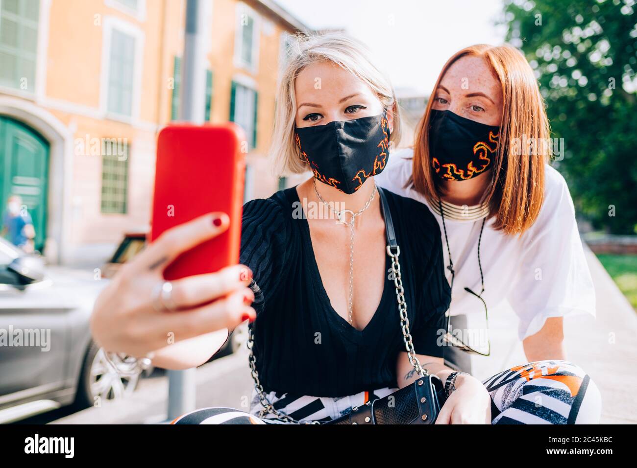 Due giovani donne che indossano maschere di fronte durante il virus Corona, seduta su una riva del fiume, prendendo selfie. Foto Stock